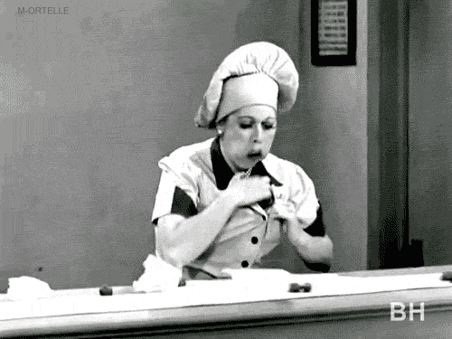 Lucille Ball, mouth full of chocolates, stuffs even more chocolates from the conveyor belt in her top.
