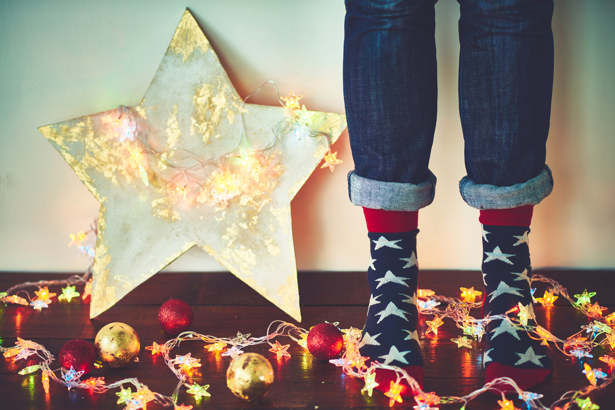 Human feet in socks with foam stars, plywood star, Christmas lights and ornaments on hardwood floor.