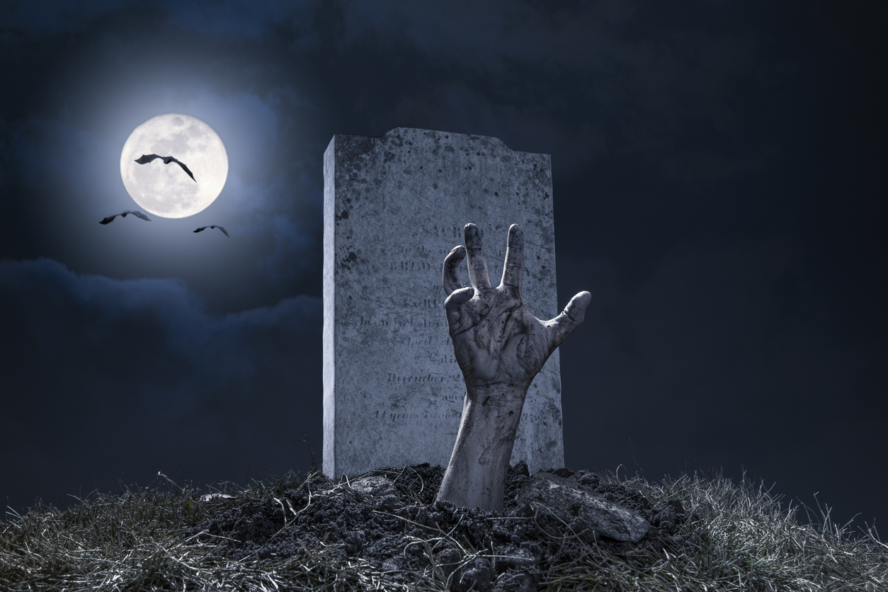  A zombie hand bursting through the grave in a graveyard on Halloween night under a full moon with vampire bats.
