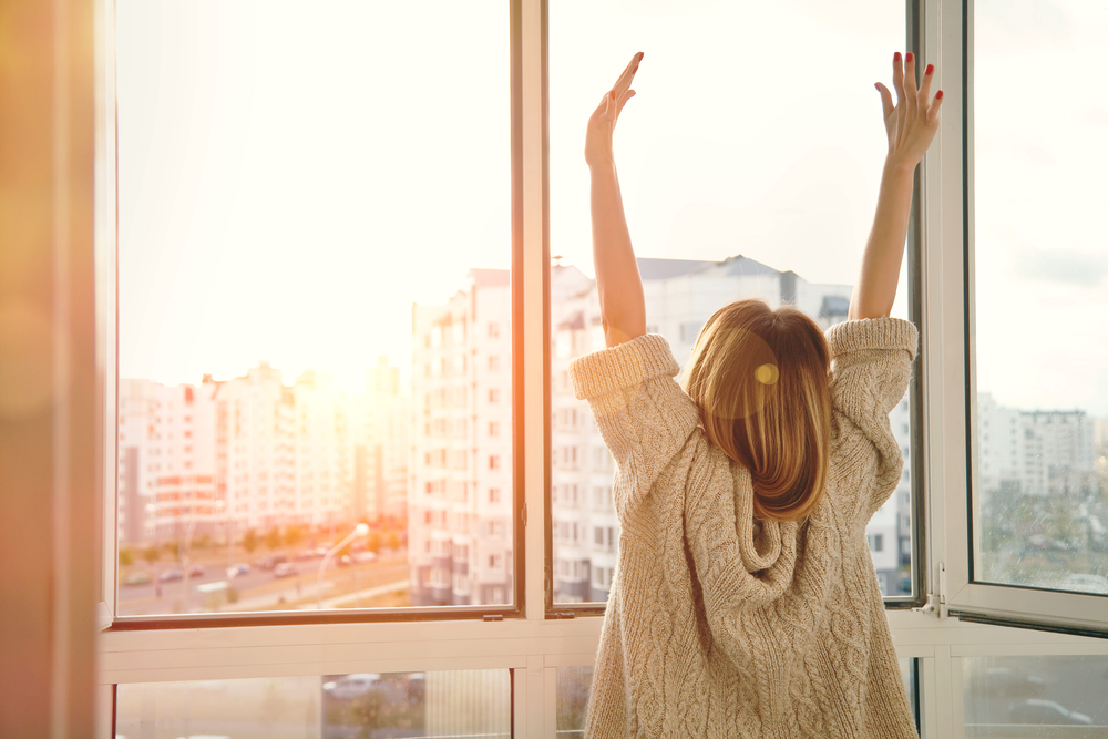 A woman, stretches in front of a window as she takes in a beautiful morning.