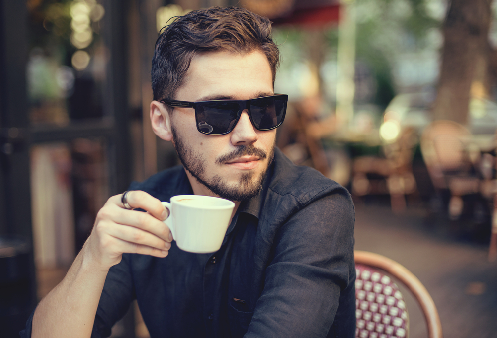 A handsome man at a cafe wears sunglasses as he sips on his coffee.