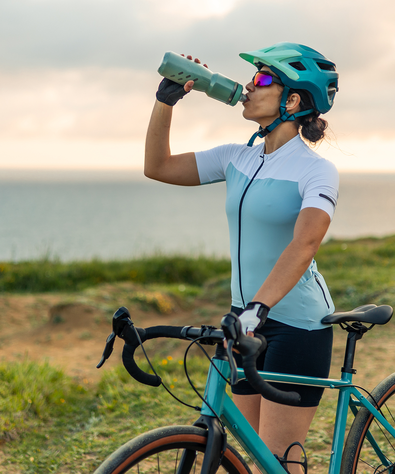 Woman drinking from a reusable water bottle beside her bike on the beach.